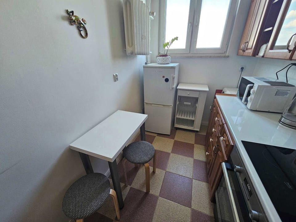A cozy breakfast nook within the kitchen features a compact white table paired with matching stools against a light wall. A refrigerator stands neatly in the corner beneath a large window that brightens the entire cooking area.
