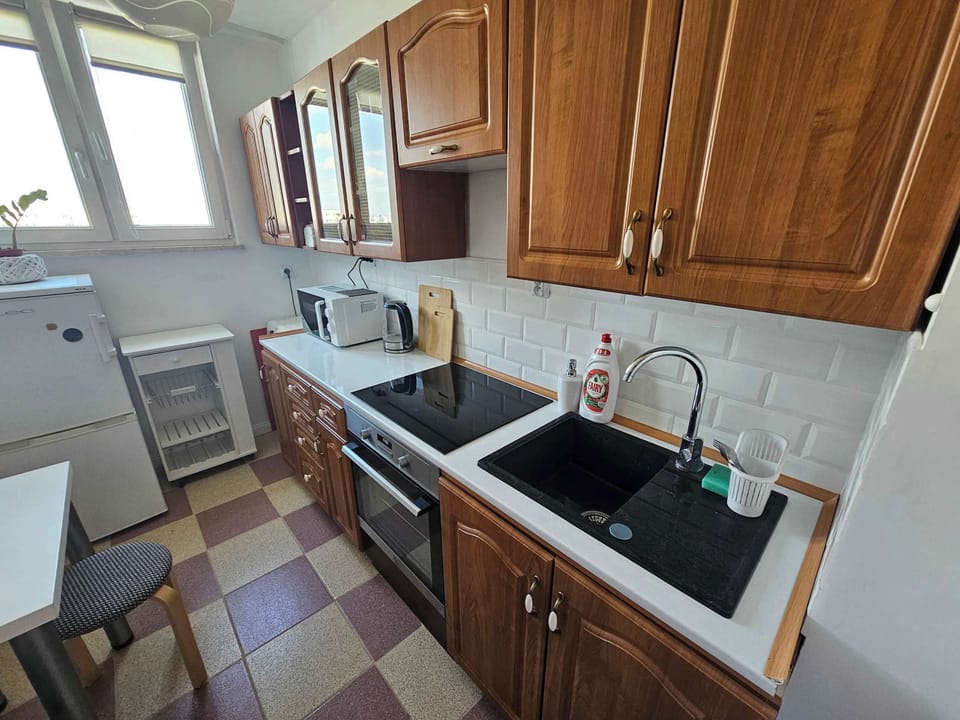 A fully equipped kitchen area showcases functional wooden cabinetry, a dark built-in sink, and a modern stovetop. Bright natural light streams through the windows, highlighting the clean tiled splashback and countertops.