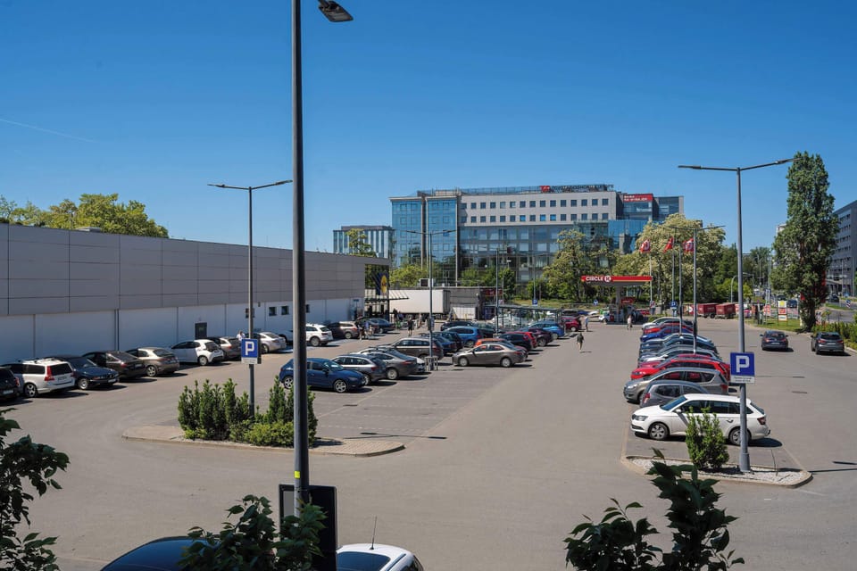 A balcony view overlooking nearby buildings, parked cars, and greenery creates a bright outdoor atmosphere. Large windows allow plenty of natural light into the apartment.