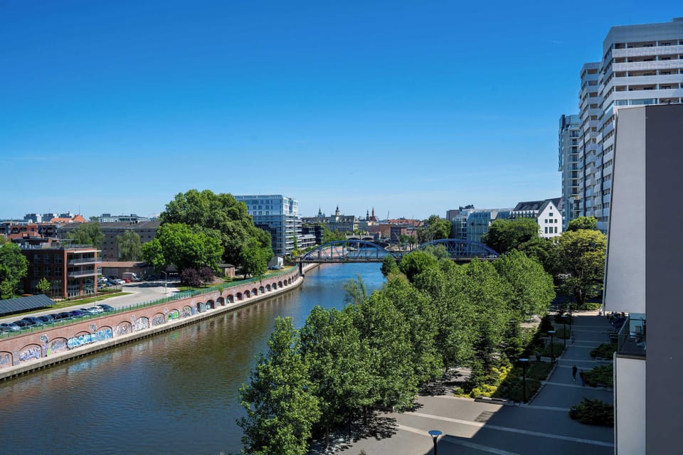 A wide view of a canal or river bordered by modern buildings and trees. The scene feels calm and connected to the surrounding city environment.