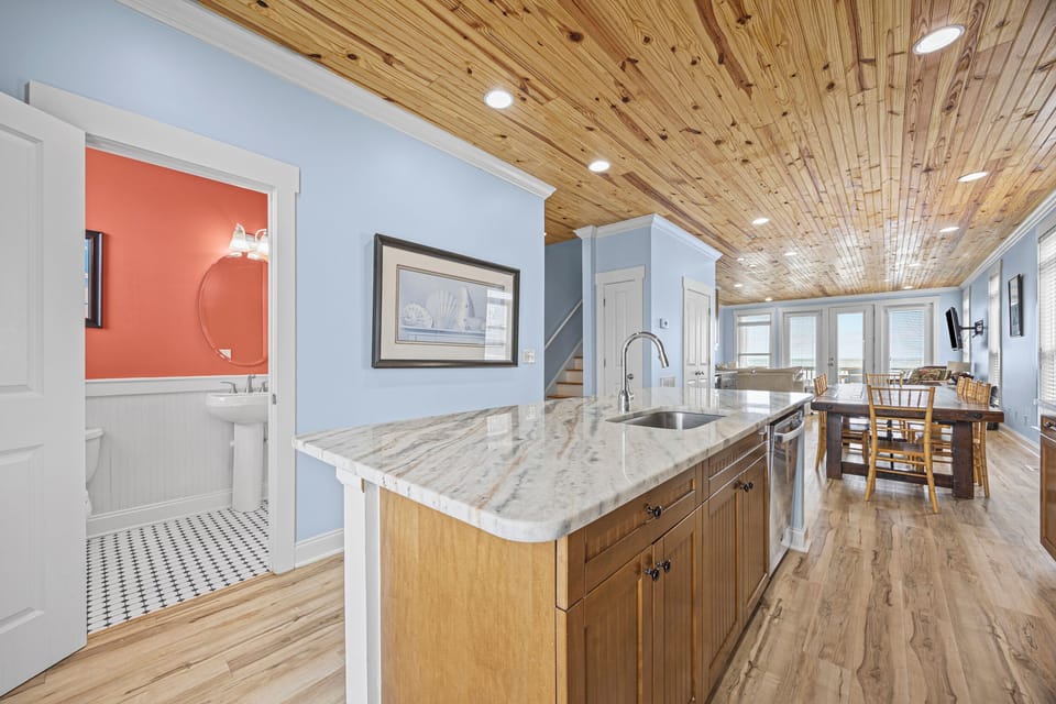 Picture yourself cooking in this airy open-concept space with its striking wood-plank ceiling and marble-topped island. Natural light floods through large windows, connecting the kitchen to a cozy dining area with a nearby bathroom for convenience.