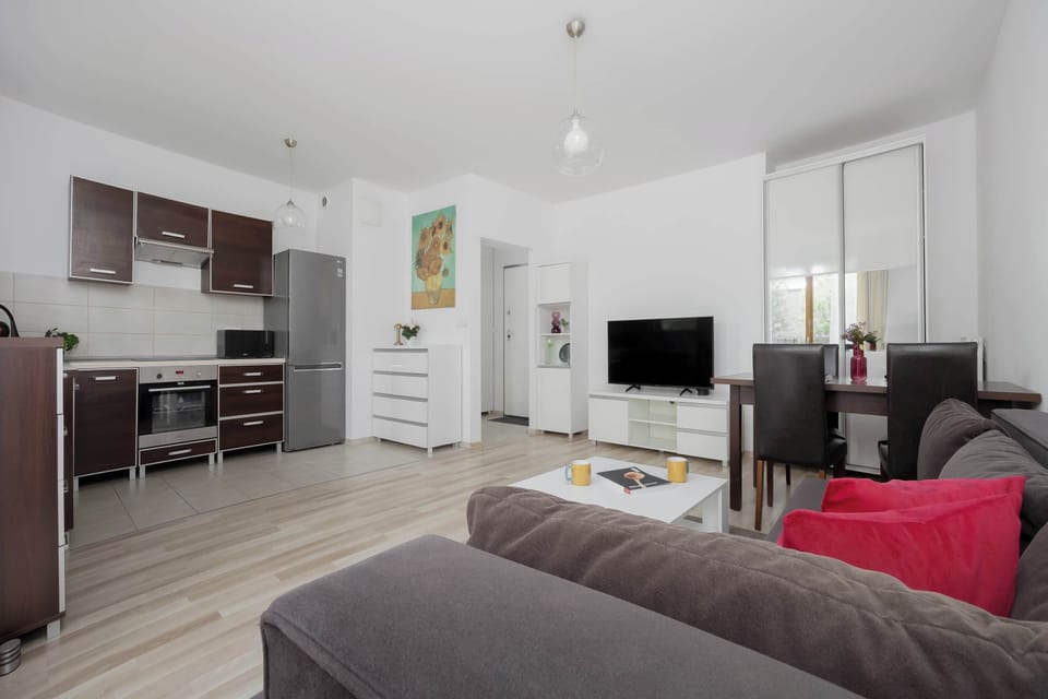An open-plan apartment interior combining the living room, dining space, and kitchen in one bright area. Large windows and pale flooring make the apartment feel spacious.