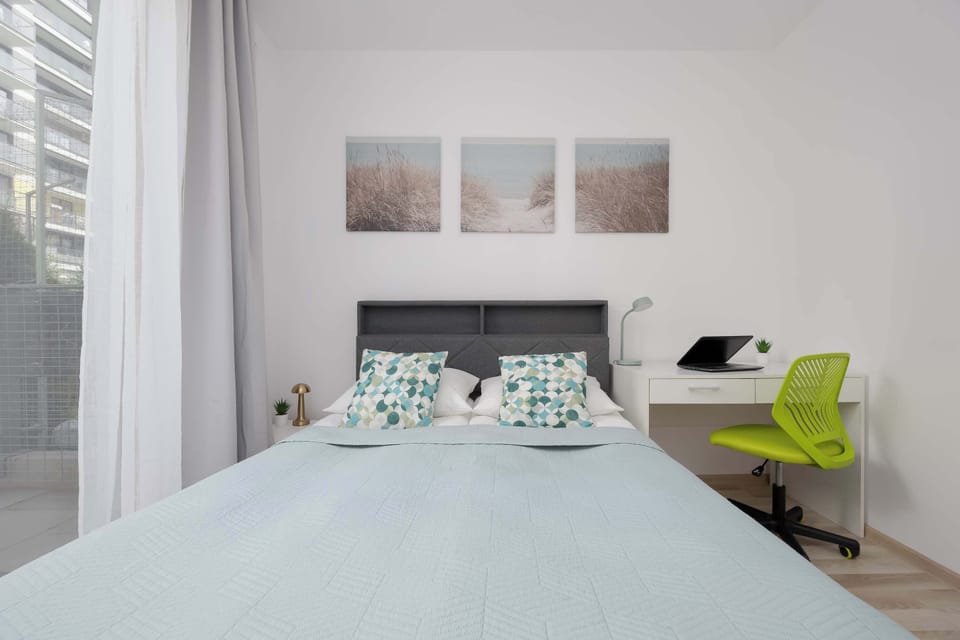 A minimalist bedroom featuring a neatly arranged bed placed beside wide windows and soft curtains. White walls and subtle décor contribute to the calm atmosphere.