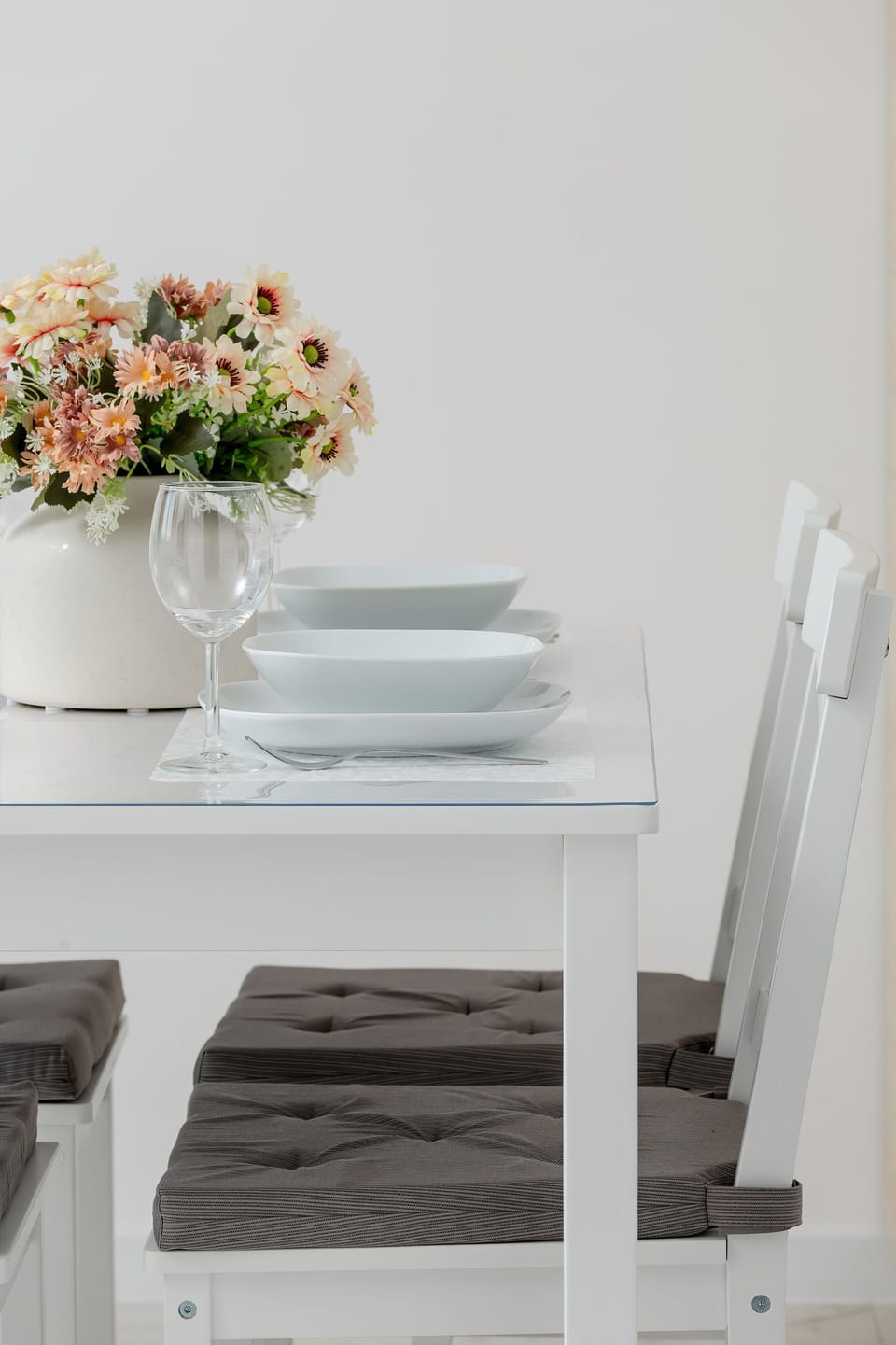 A close-up shot focuses on the dining table setup, highlighting white ceramic plates stacked on fabric placemats. A bouquet of artificial flowers adds a soft touch of color next to the white wooden chairs.