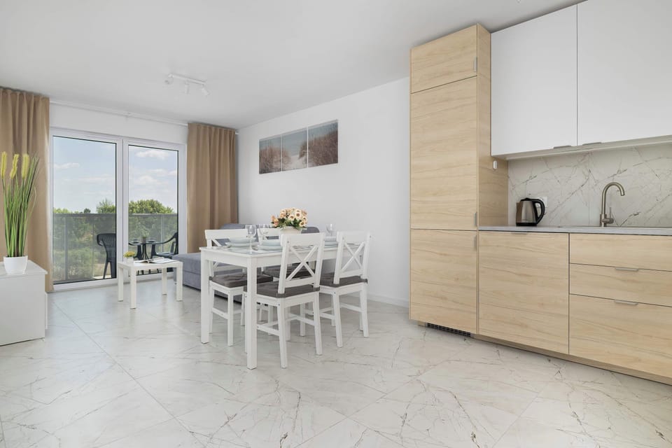 This wide angle captures how the open-plan layout flows directly from the wooden-faced kitchen cabinetry into the dining zone. Sun streams in from the large balcony doors at the back of the room.