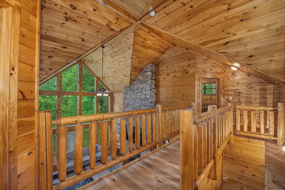 Natural light fills this lofted space through floor-to-ceiling windows, revealing rich knotty pine walls and vaulted ceilings. The handcrafted log railings and stone fireplace add rustic mountain charm throughout.