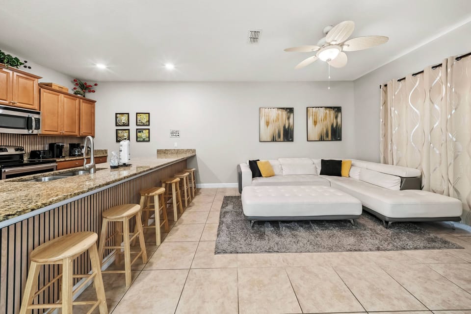Relax in the spacious living area featuring a stylish sectional sofa and vibrant yellow accents. The warm light wood cabinetry and tiled floors make it the perfect spot to unwind and share memorable moments with your loved ones.