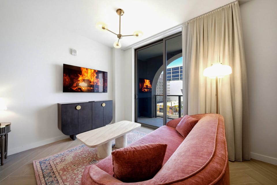 Enjoy the cozy charm of this living area with its inviting pink sofa, warm chandelier light, and elegant decor. The floor-to-ceiling windows provide a stunning glimpse of the vibrant outdoor view, perfect for relaxation.