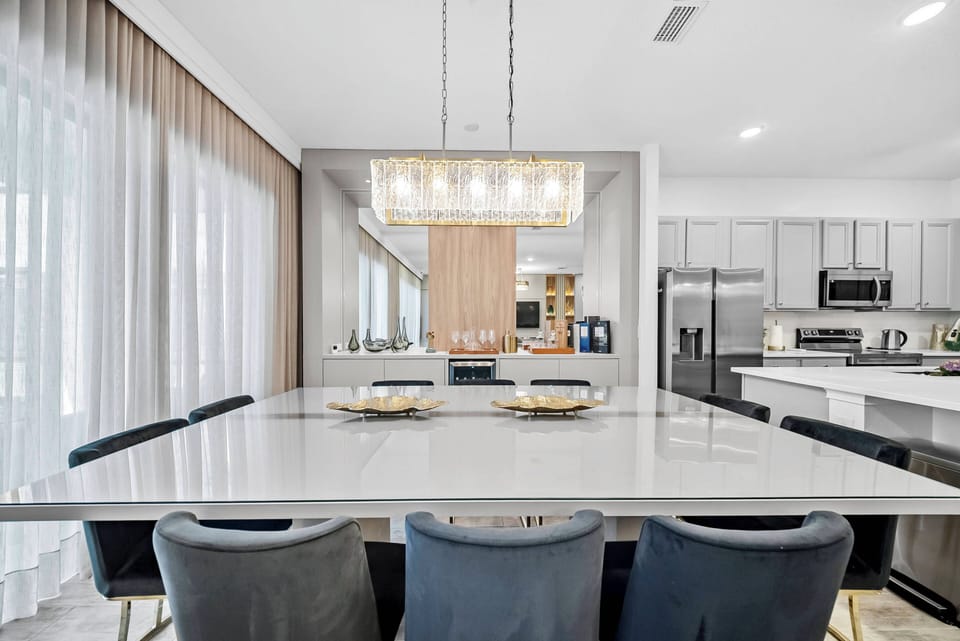 Experience a chic dining area featuring a sleek white quartz table, perfect for gatherings beneath a modern chandelier. The soft taupe curtains filter natural light, creating an inviting ambiance in this contemporary space.