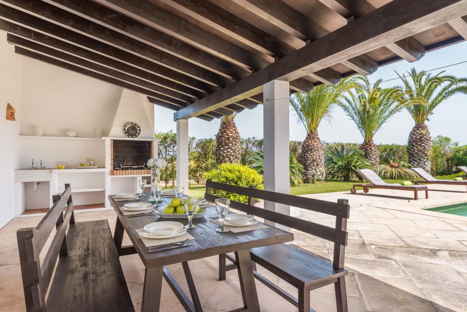 Mesa comedor exterior bajo porche de madera, con barbacoa y vistas a la piscina 