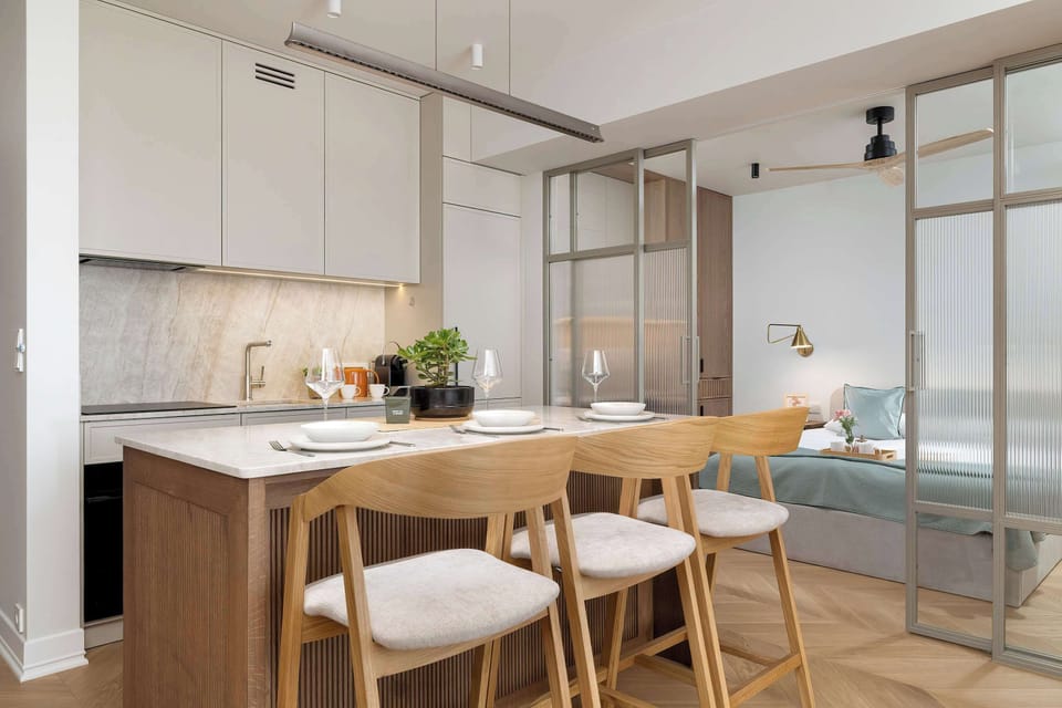 A dining area with a wooden table and upholstered chairs is positioned next to the kitchen. Glass partitions and modern lighting enhance the bright and contemporary character of the space.