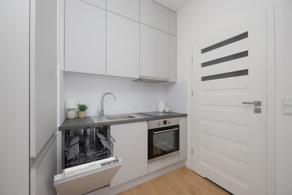 A minimalist kitchenette with white cabinetry, built-in appliances, and compact wooden flooring. Clean surfaces and subtle lighting emphasize the modern design.