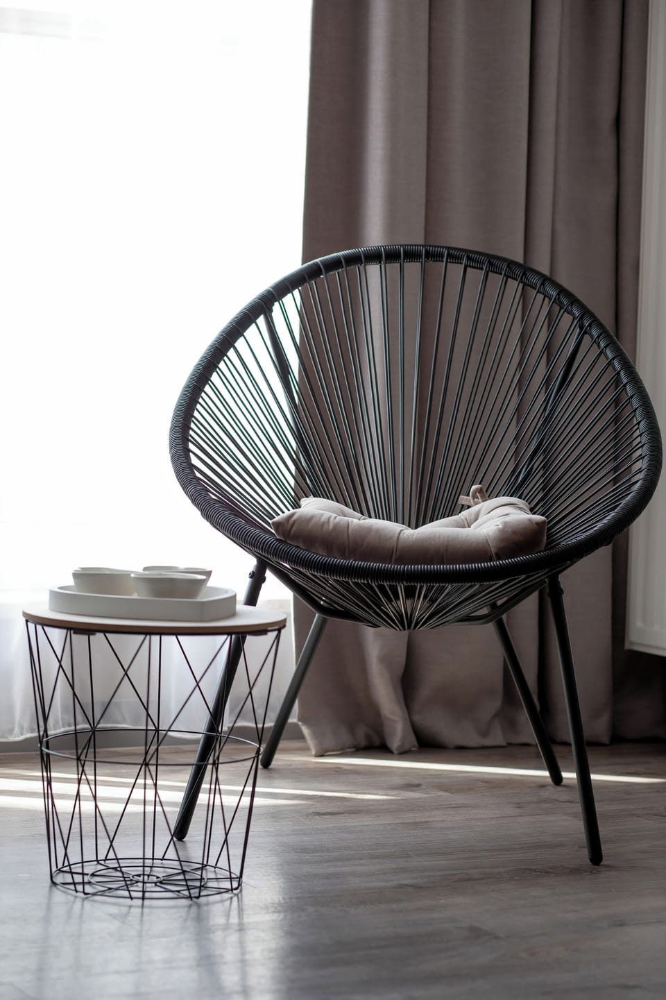 A close-up, vertical shot isolates one of the black woven chairs paired with a small geometric accent table. The sheer curtain in the background diffuses the outdoor light softly.