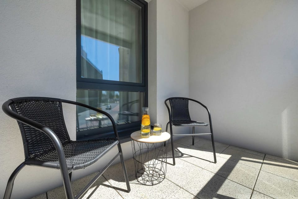 This close-up angle of the balcony highlights the two black woven armchairs catching the bright afternoon sun. A small table between them holds a bottle and a glass, offering a relaxing spot to unwind.