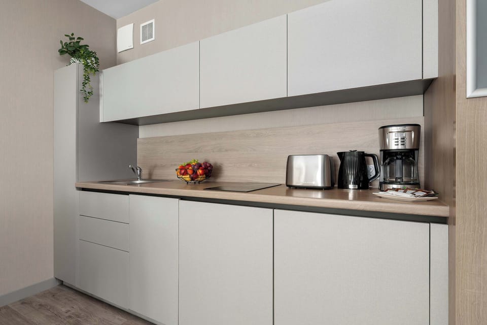 A straight-line kitchenette features minimalist light grey cabinets and a matching wood-textured backsplash. The countertops are clean, creating a streamlined and modern cooking zone.