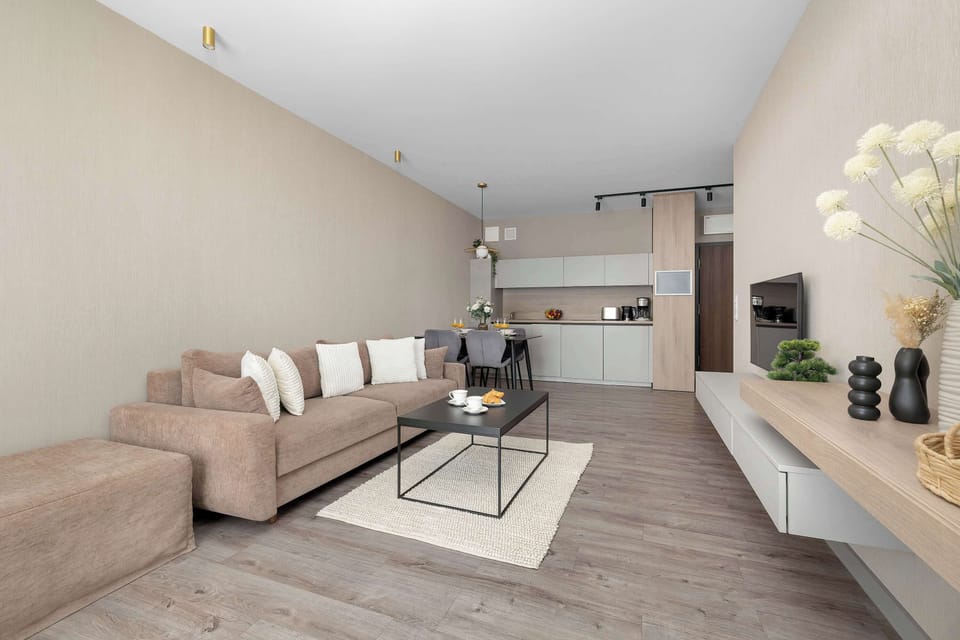 Looking across the entire apartment, the spacious layout unifies the lounging area, dining setup, and kitchen. The warm, sandy color palette creates a cohesive and inviting environment throughout.