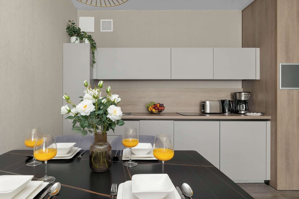A dining table set for guests is positioned in front of the kitchenette, accented by a prominent bouquet of white roses. Glasses containing orange juice stand ready alongside neat stacks of square white plates.