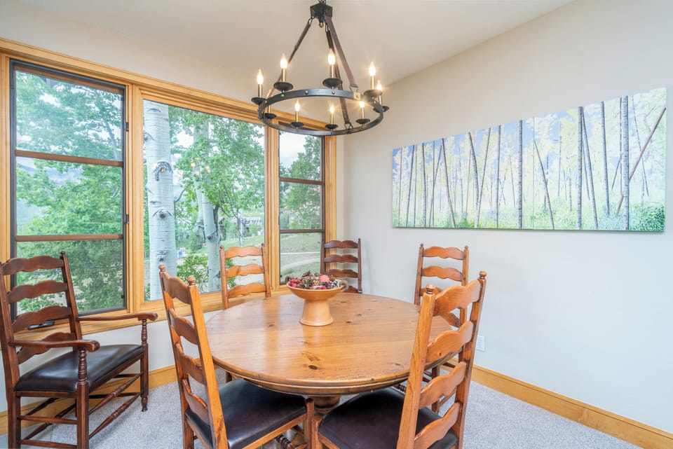 Gather around the handcrafted wood table under a rustic chandelier, with natural light streaming through windows overlooking serene aspen trees—your perfect space for memorable meals together.