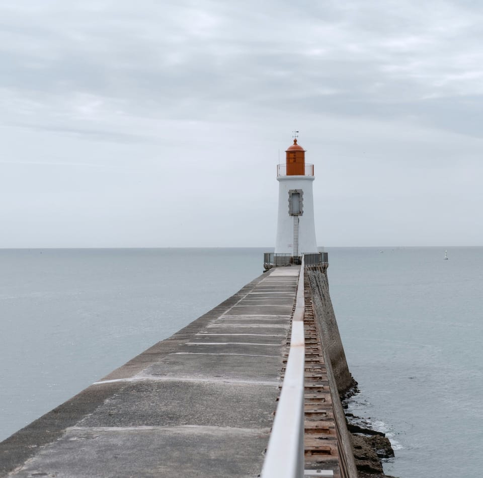 A solitary red-topped lighthouse at the end of a narrow concrete jetty under an overcast grey sky.