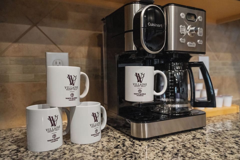 Coffee station with drip coffee maker, villain-themed mugs, and black counter.