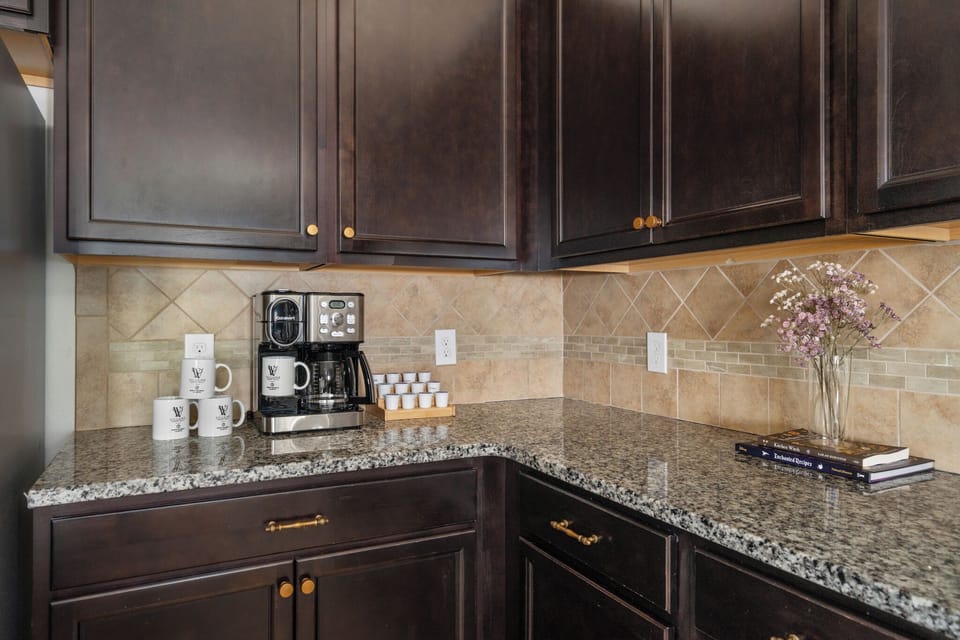Kitchen counter with sink, coffee supplies, dark cabinets, and tile backsplash.