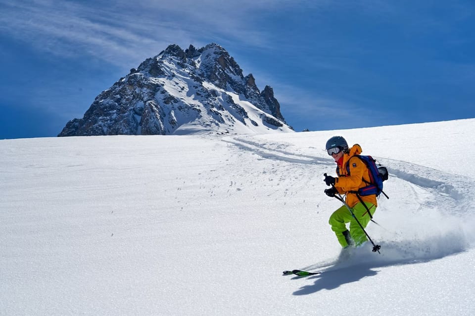 A skier carving through fresh powder snow with a dramatic Alpine peak in the background.