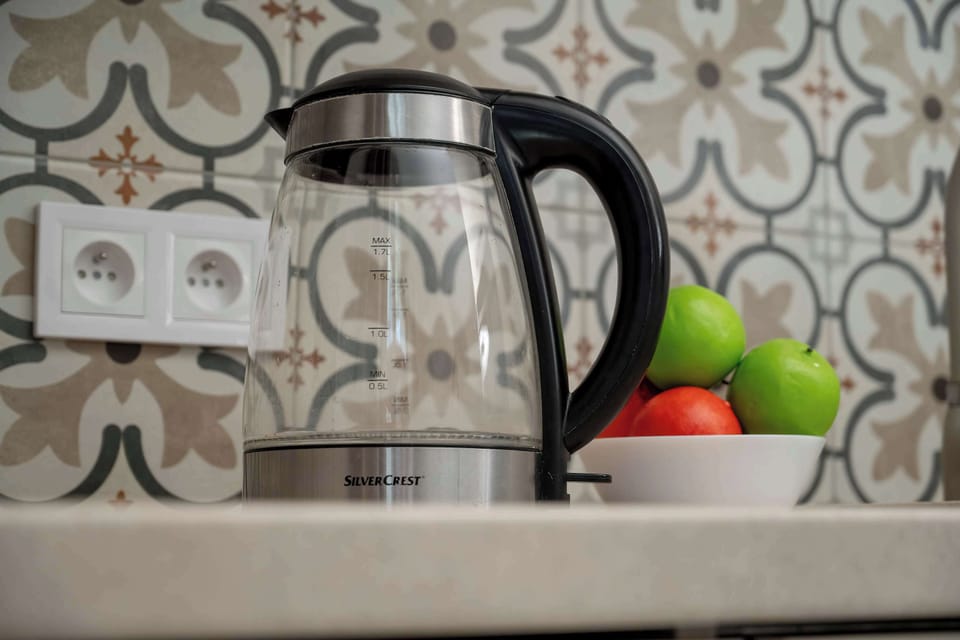 A close-up of a kitchen countertop featuring an electric kettle and fruit placed against decorative patterned tiles. Small details add warmth to the compact space.