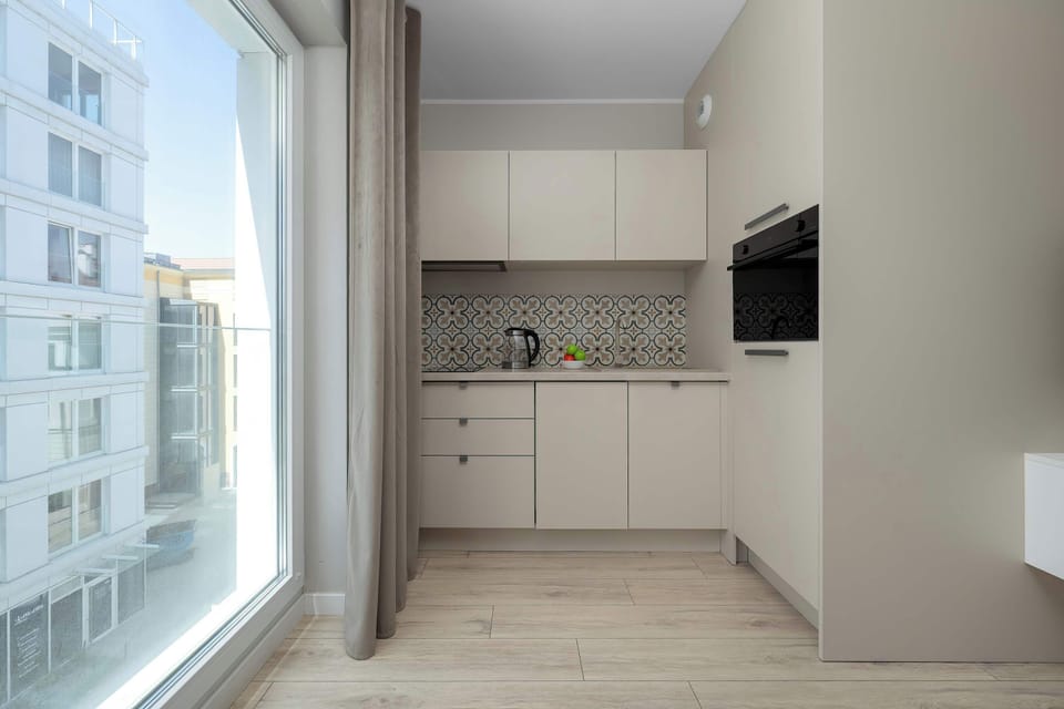 A kitchen area positioned beside large windows, allowing abundant daylight to enter the room. White cabinets and patterned backsplash details add a modern touch.