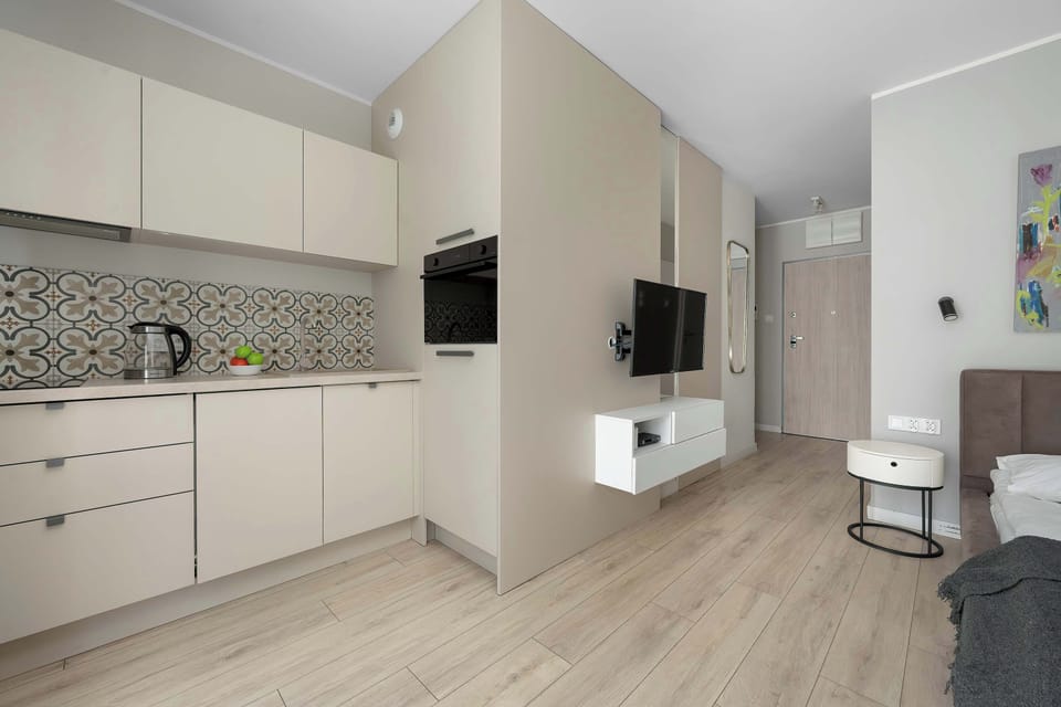A wide view of the studio apartment showing the sleeping area, television, and kitchenette integrated into one functional layout. Bright finishes make the room appear larger.