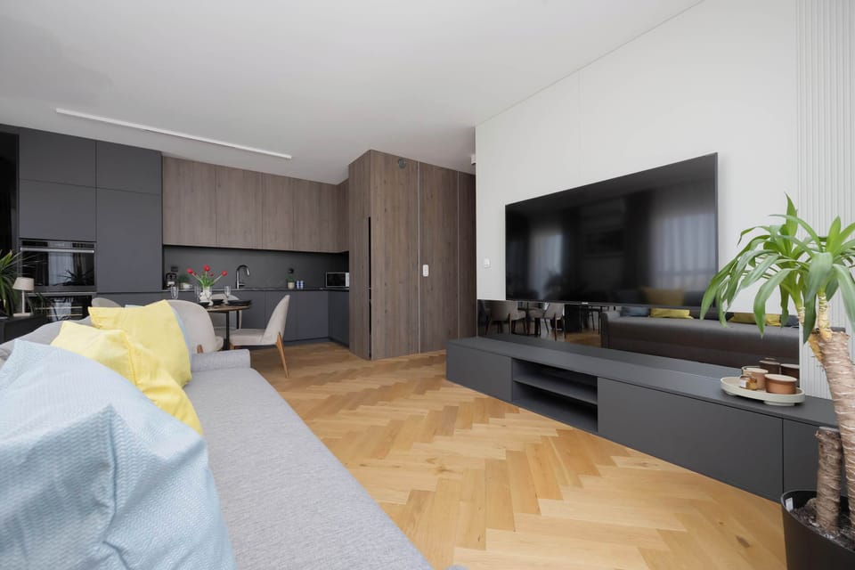 A modern living room with a large TV and a comfortable bed visible in the foreground. The space is bright and decorated in neutral colors.
