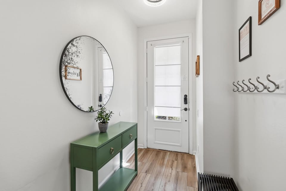 Entrance hallway with wood-look flooring, green console table, round distressed mirror, and white front door.