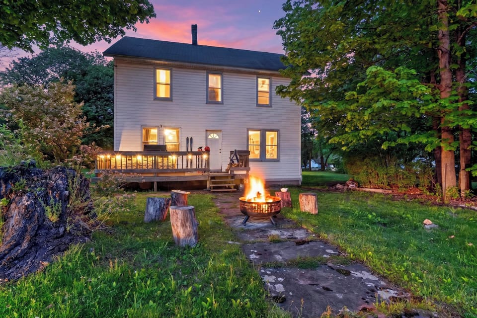 Twilight backyard retreat with a glowing fire pit, deck seating, and peaceful wooded charm.