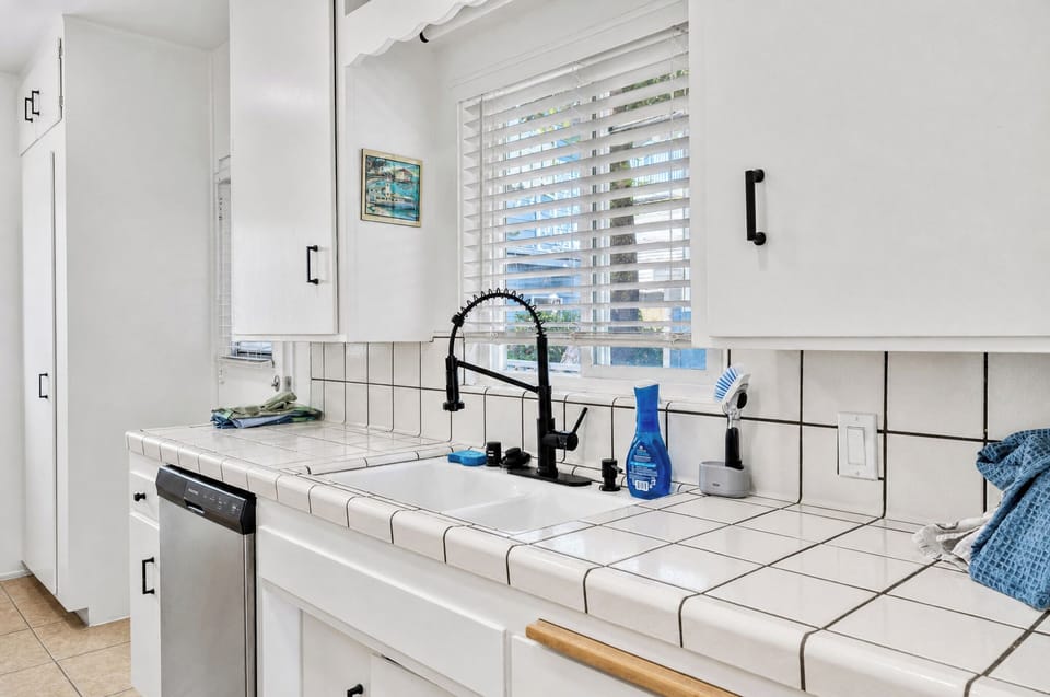 Your modern kitchen features tile countertops, a sleek black faucet, and plantation shutters that invite natural light—a bright, functional space for preparing meals during your stay.