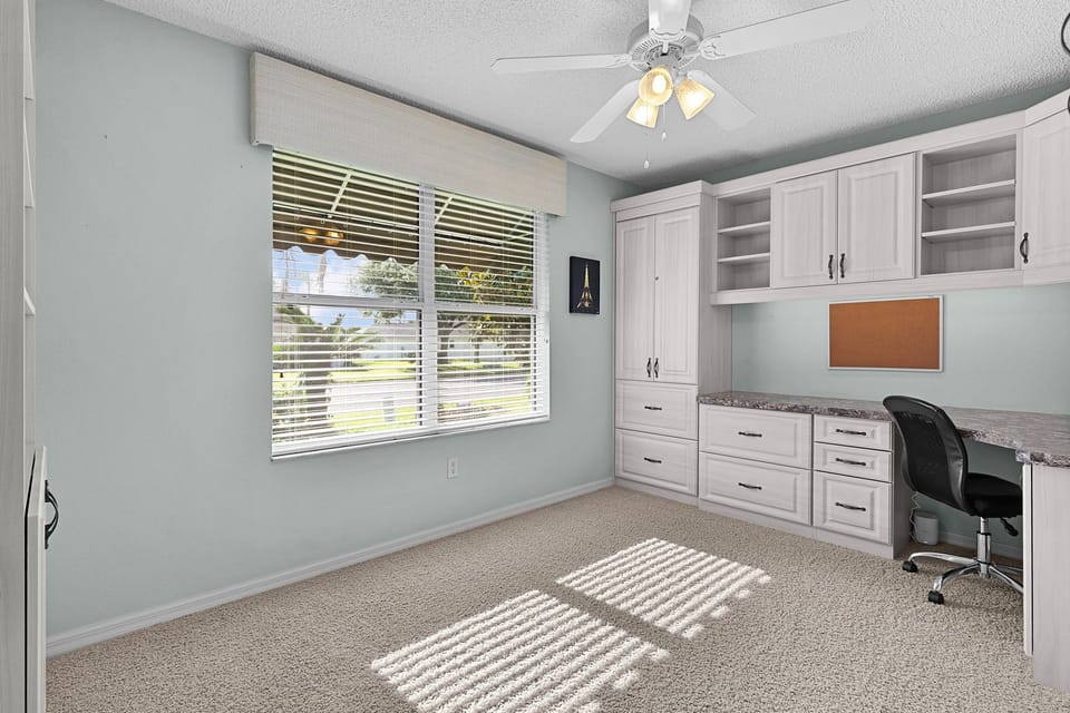 Step into a bright home office with custom cabinetry, a spacious desk with garden views, and soft carpeting underfoot. Your productive retreat awaits with built-in storage and calming blue walls.