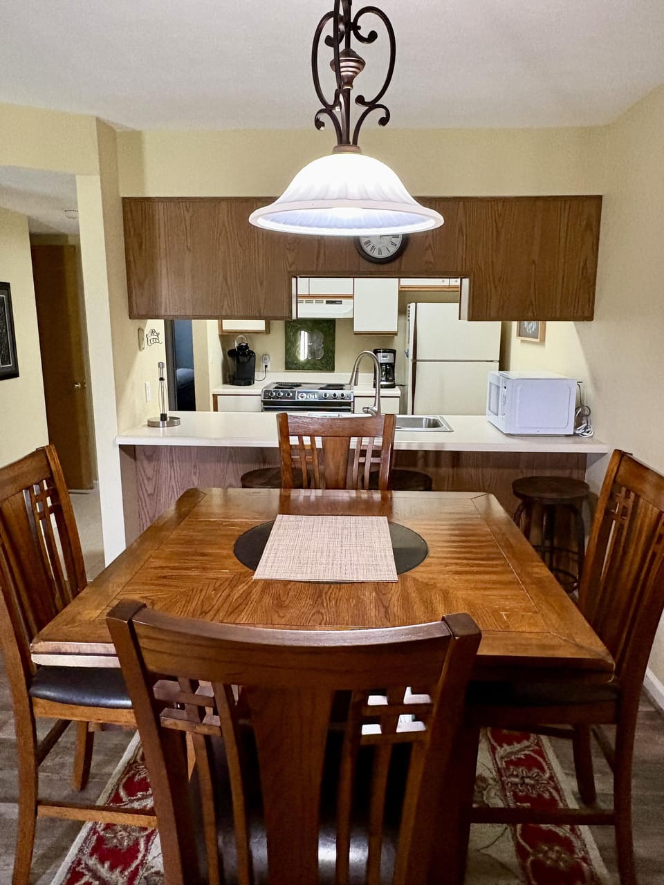 Wooden dining table with four chairs sits on patterned rug beneath a pendant light over the kitchen pass-through.