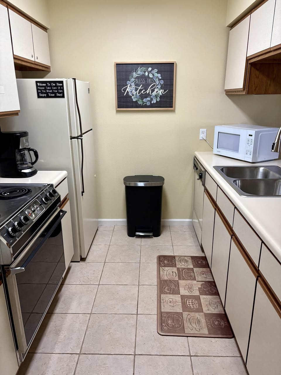 Galley kitchen with beige tile floor, white appliances, wood-trimmed cabinets, and coffee-themed floor mat.