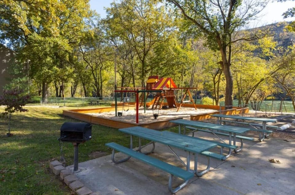 Picnic area with green wooden tables and a charcoal grill near a children's playground in a leafy park.
