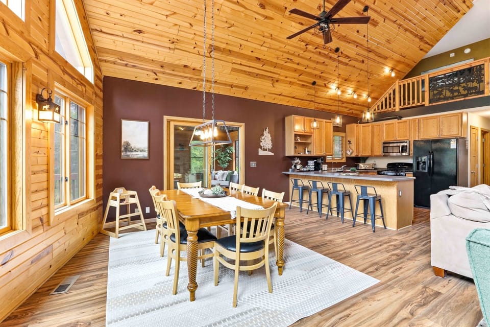 Expansive open-concept dining and living area with vaulted wood ceilings, warm finishes, and a seamless flow to the kitchen and fireplace lounge.