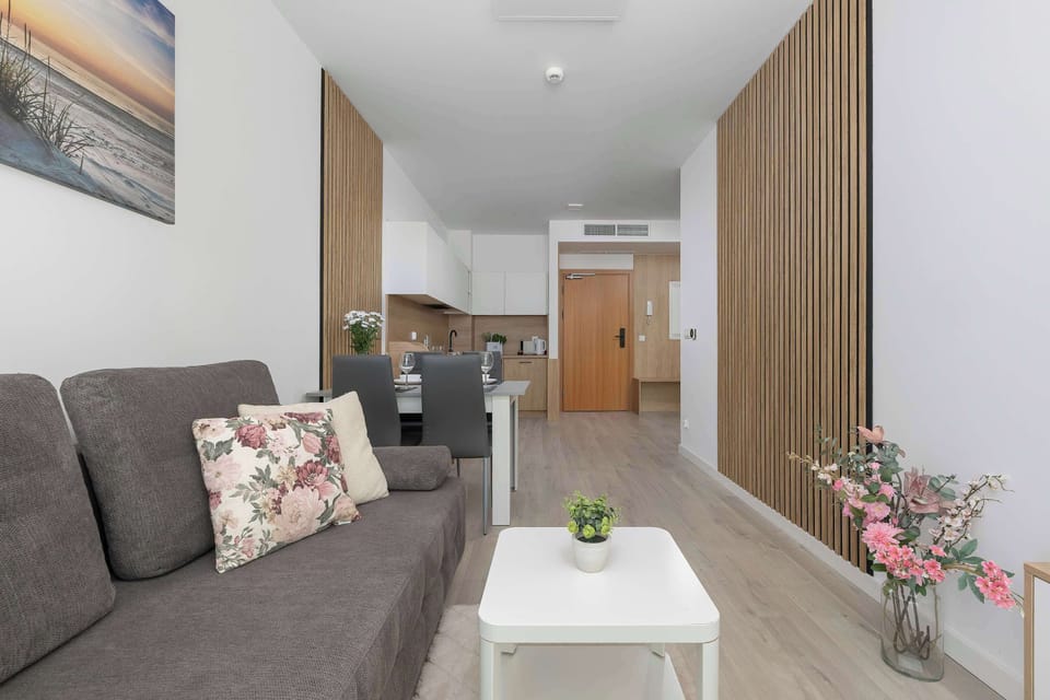 A view of the living room showing a gray sofa siand a coffee table. The interior is bright, well-lit, and finished with natural wood elements.