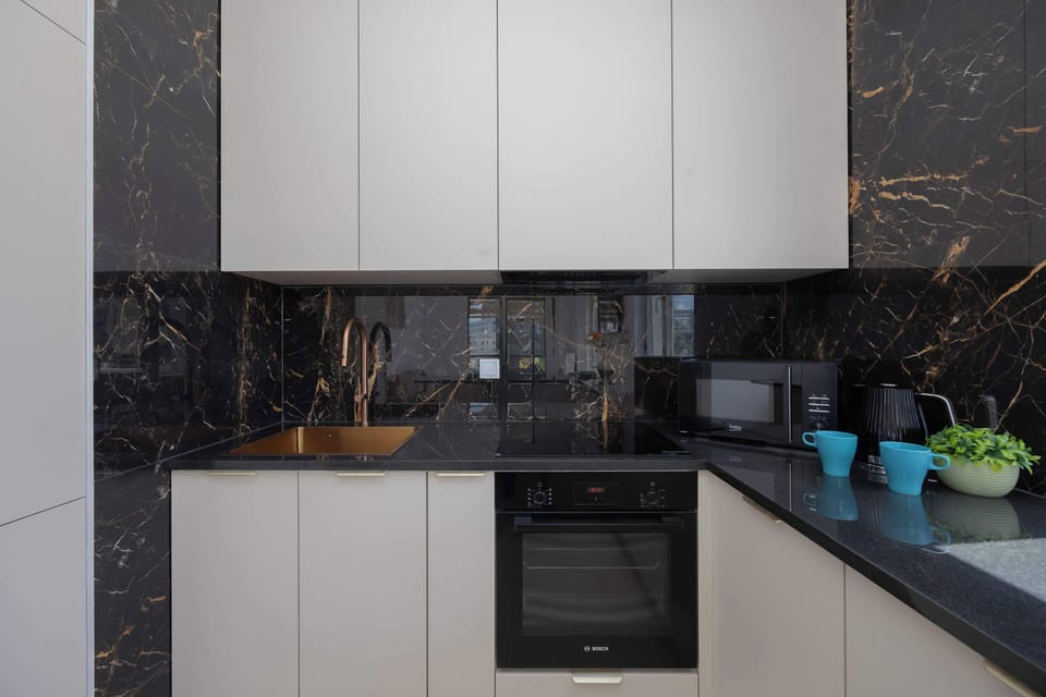 A sleek, minimalist kitchenette showcases glossy white upper cabinets and a dark, marbled backsplash. A black built-in oven and induction hob are integrated into the countertops, accompanied by a black electric kettle.