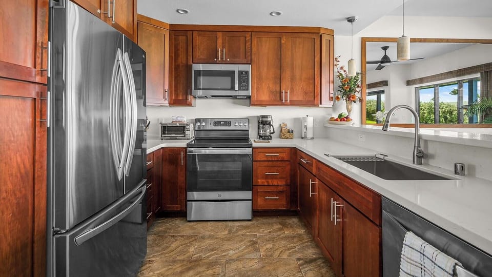 Your gourmet kitchen awaits with sleek stainless steel appliances, rich wood cabinetry, and abundant counter space—perfect for preparing island-inspired meals while enjoying garden views through the window.