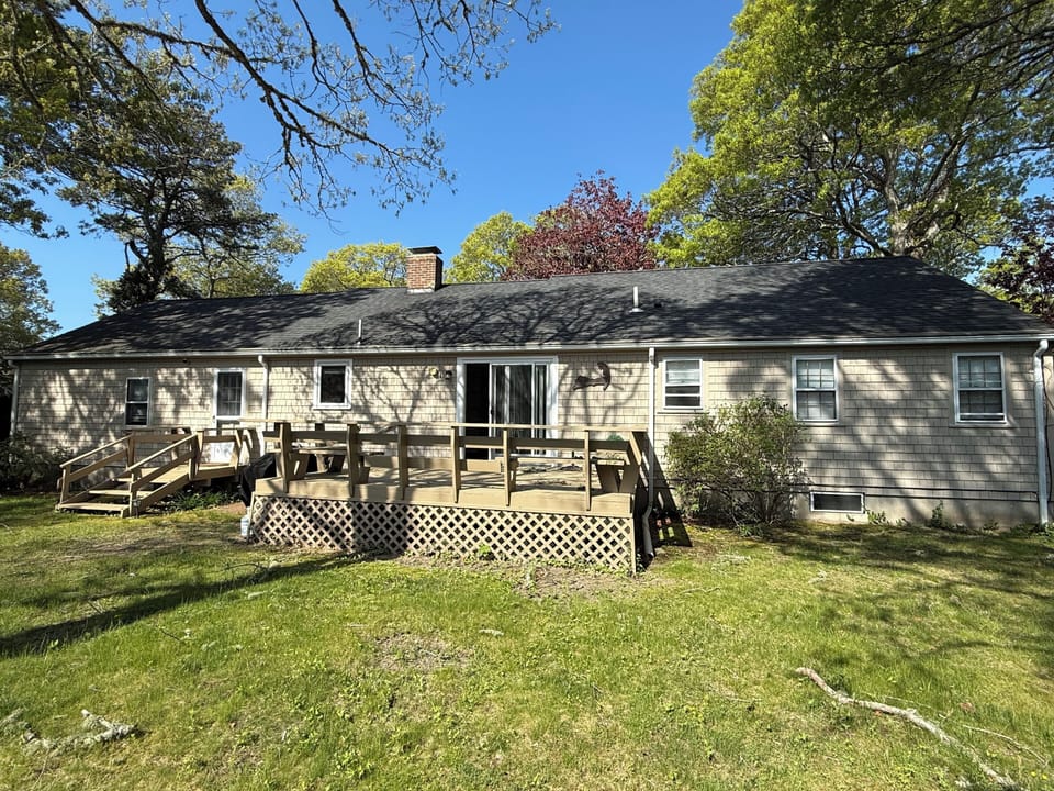 A charming single-story vacation home features a spacious wooden deck with outdoor seating, perfect for relaxing under the shade of mature trees in this peaceful, tree-lined setting.