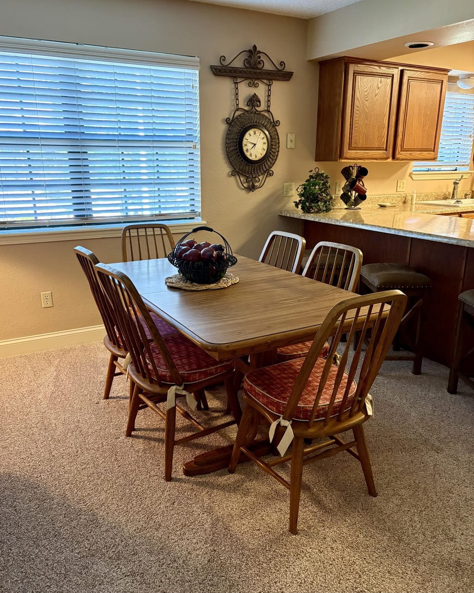 Dining area features a wood table with six spindle-back chairs on plush beige carpet next to granite kitchen counters.