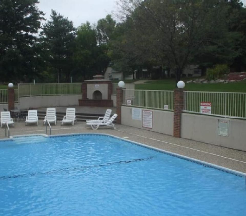 Outdoor pool with clear blue water, white lounge chairs on textured concrete deck, and brick pillars with globe lights.