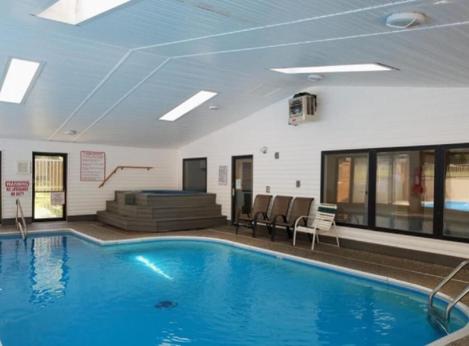 Indoor pool with curved edges, textured concrete deck, stacked lounge chairs, and skylights above.