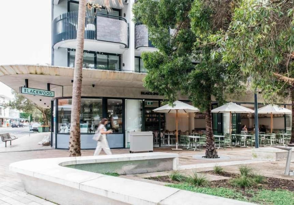 Enjoy outdoor dining at Blackwood Pantry, a vibrant corner cafe featuring inviting outdoor seating, patio umbrellas, modern architecture, and surrounding greenery.