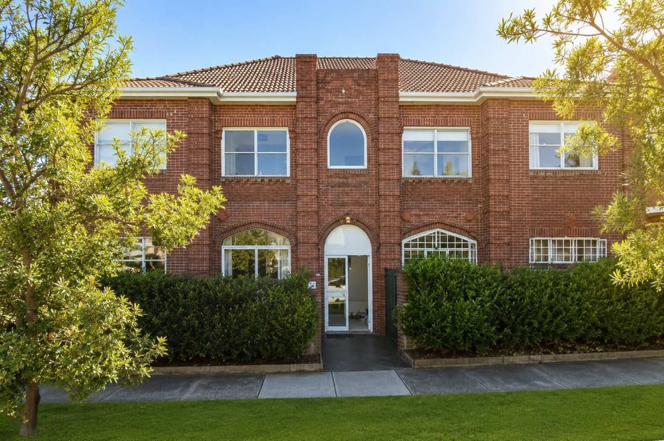 Welcome to this classic red brick apartment building, surrounded by lush green hedging, manicured lawns, and mature trees creating an inviting curb appeal.