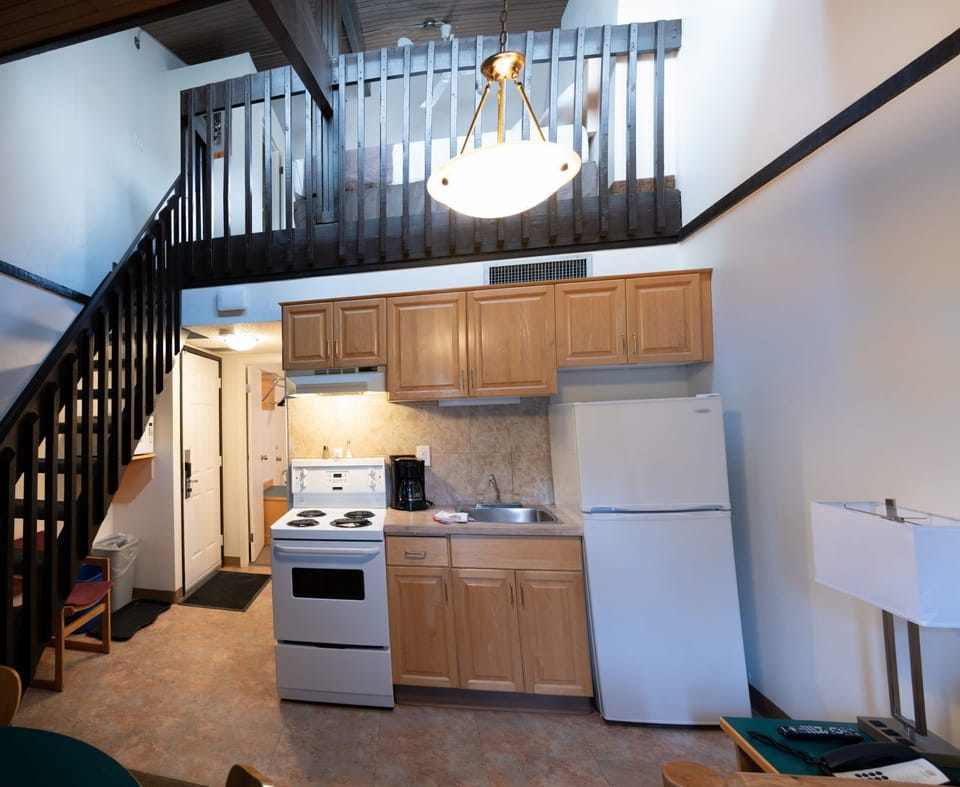 Open kitchen with light wood cabinets, white stove, fridge, and coffee maker — stairs to loft just behind