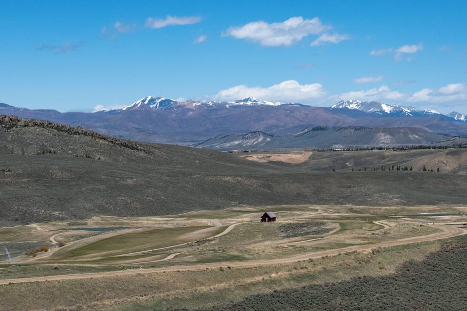 Expansive mountain valley landscape featuring snow-capped peaks, rolling hills, and a solitary barn structure in the distance.