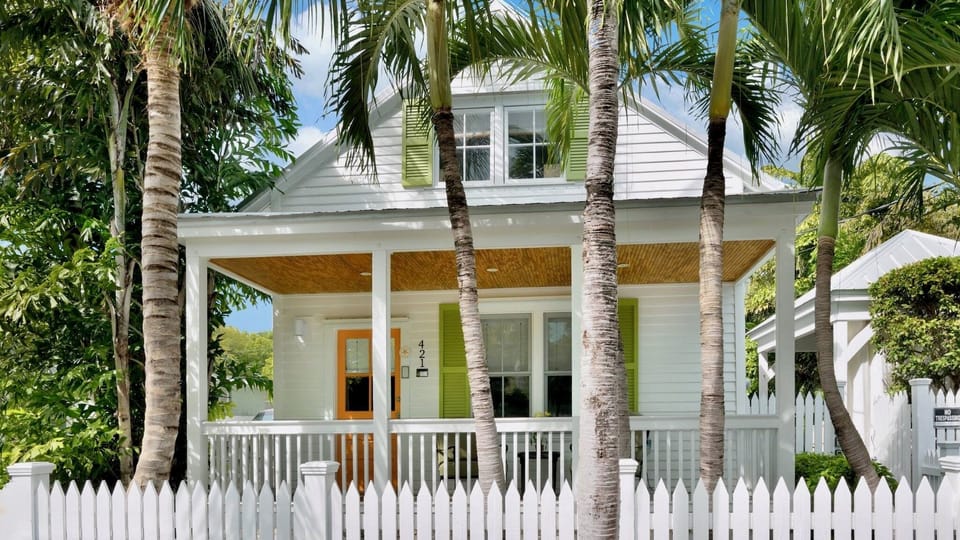 A charming coastal cottage with tropical palms and fresh green shutters marks your arrival, nestled in a lush, private neighborhood under sunny skies.